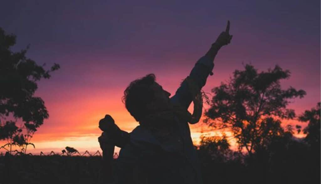 A man silhouetted against the sunset points toward the sky